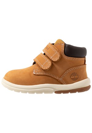 Botas de ante color arena con dos correas de velcro, detalle de cuero marrón en la parte superior, suela de goma blanca con dibujo en marrón claro y logo en el lateral.