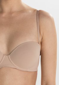 Beige, polstret bh med sømløse kopper og tynne justerbare stropper med subtil merkevare. Jevn stofftekstur som gir et minimalistisk design.