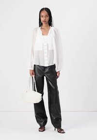 Blusa bianca trasparente con dettagli a bottoni, abbinata a pantaloni larghi in pelle nera e sandali neri con cinghie. Tiene una borsa bianca.