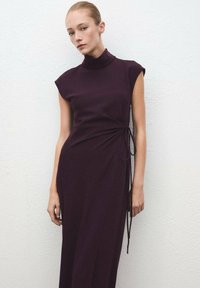Mujer con el pelo recogido que lleva un vestido sin mangas de cuello alto, de color púrpura oscuro, ajustado en la cintura, de pie contra una pared blanca con textura.
