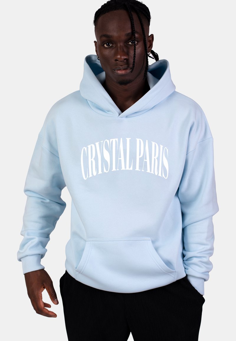 Felpa blu chiaro con tasca frontale e testo bianco "CRYSTAL PARIS". Caratterizzata da un cappuccio con coulisse e polsini a costine. Indossata con pantaloni neri.