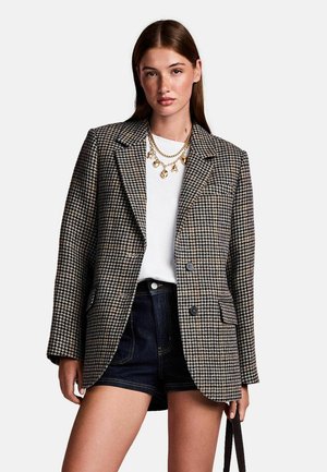 Houndstooth-patterned blazer in black and beige, featuring a notched lapel, two buttons, and a textured fabric. Paired with a white top and denim shorts.