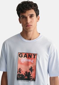 Camiseta de algodón azul claro con un estampado gráfico de palmeras en coral y negro, con el texto “GANT Resort” y “American Sportswear Since 1949.”