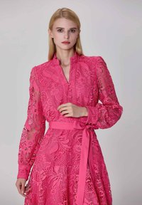 Femme blonde portant une robe en dentelle rose avec des motifs floraux, un col montant, des manches bouffantes et une ceinture nouée à la taille, se tenant devant un fond neutre.