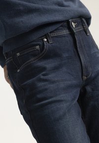 Jeans de mezclilla azul oscuro con corte slim, que presentan costuras en contraste, bolsillos delanteros y un cierre de botón metálico en la cintura.