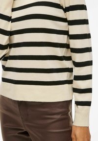 Pull beige avec des rayures horizontalement noires, poignets et ourlet côtelés ; accompagné d'un pantalon marron à texture lisse, vue focalisée sur le côté.