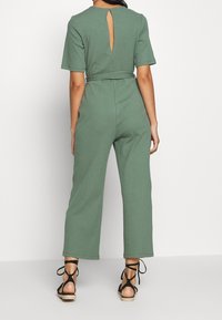 Groene geribde jumpsuit met korte mouwen, opening met sleutelhals aan de achterkant, een taille met riem en wijde broekspijpen. Gecombineerd met zwarte vetersandalen.
