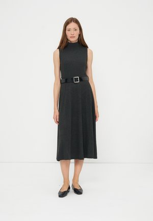 Femme portant une robe midi sans manches gris foncé avec ceinture noire et chaussures plates noires, debout devant un fond clair uni.