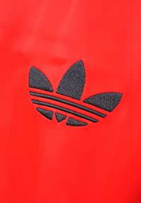 Rdeča tekstilna podlaga z črnim vezenim logotipom Adidas, ki ima tri listnate oblike in tri vodoravne črte pod njimi.