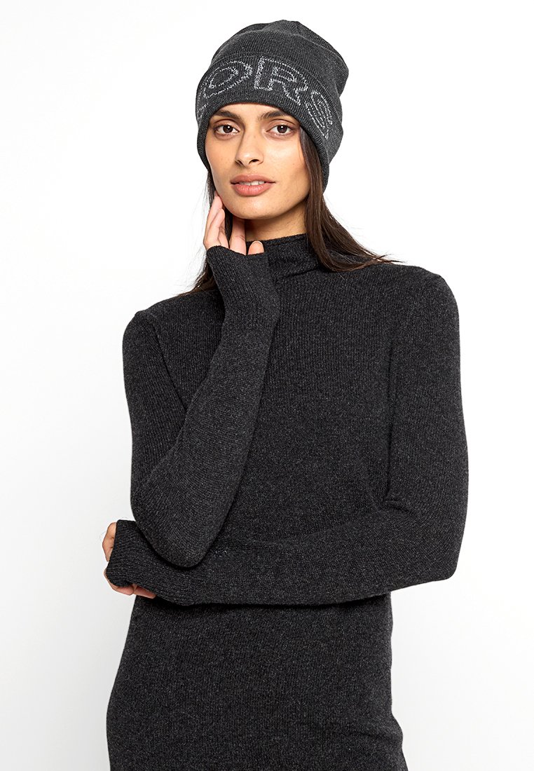 Donna che indossa un maglione a collo alto in maglia grigio scuro e un cappello abbinato con la scritta "ORS", mentre posa con una mano vicino al viso su uno sfondo bianco.