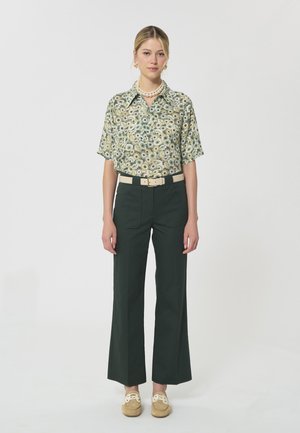Femme portant un chemisier vert à manches courtes à fleurs, un pantalon large vert foncé, une ceinture beige, des mocassins beige et un collier et des boucles d'oreilles en perles.