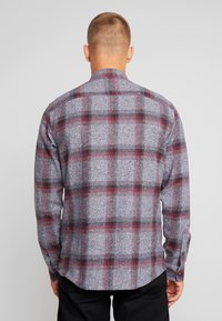 Camicia a quadri grigia con accenti rossi e neri, maniche lunghe, polsi con bottoni e colletto. La texture appare morbida e tessuta.
