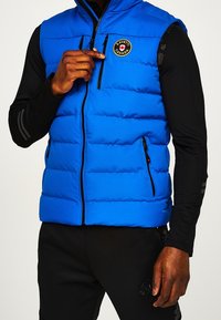 Blauer Steppweste mit horizontaler Quiltung, Reißverschluss vorne und Taschen, mit einem runden Logo auf der Brust, kombiniert mit einem schwarzen Langarmoberteil.