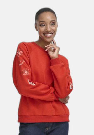 Femme souriante portant un sweat-shirt rouge avec des symboles blancs de soleil, œil, cœur et yin-yang sur les manches, posant avec la main près du visage.