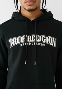 Čierna mikina s kapucňou s bielym logom a textom "TRUE RELIGION BRAND JEANS" a so sťahovacími šnúrkami a kovovými koncami. Hladká textúra látky, ležérny dizajn.