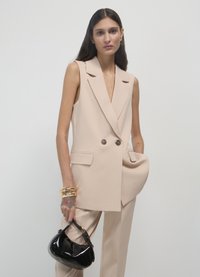 Blazer beige senza maniche dal taglio sartoriale con due bottoni e tasche, abbinato a pantaloni beige e una piccola borsa nera in pelle verniciata.