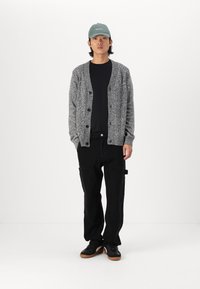 Grå strikket cardigan med knapper, båret over en svart skjorte. Svart cargo-bukser og svarte sneakers med gummisåler. Grønn caps fullfører antrekket.
