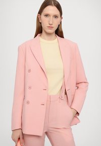 Light pink double-breasted blazer with matching trousers, paired with a pale yellow top. Smooth fabric, tailored fit, minimalistic buttons.