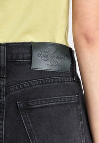 Juodi džinsai iš džinsinio audinio su lygiu paviršiumi; turi juodą odinį pleistrą su įspaustu "Calvin Klein Jeans" logotipu juosmens srityje.