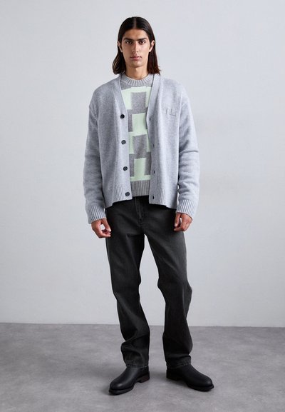 Cardigan grigio con bottoni, indossato sopra un maglione a fantasia verde e grigio. Il modello indossa jeans scuri e scarpe slip-on nere. Sfondo semplice.