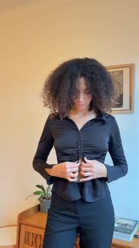 Camisa preta justa com botões, gola com colarinho, frente franzida e mangas compridas, combinada com calças pretas. Textura lisa, corte sob medida.