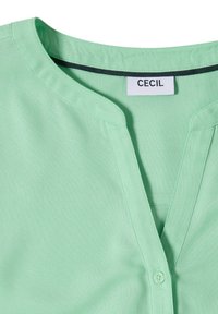 Col de blouse vert clair en V avec un petit bouton et des détails cousus, avec une étiquette blanche de la marque indiquant "CECIL."