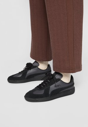 Czarne sneakers Puma z panelami z zamszu i skóry, noszone z kremowymi skarpetkami i brązowymi spodniami o szerokich nogawkach na jednolitym tle.