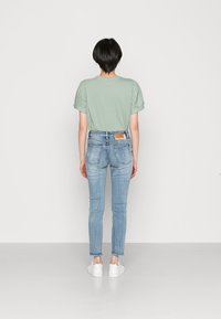 Persona con cabello corto negro, vestida con una blusa de manga abullonada verde claro, jeans de mezclilla azul claro y zapatillas blancas, de pie frente a una pared blanca.