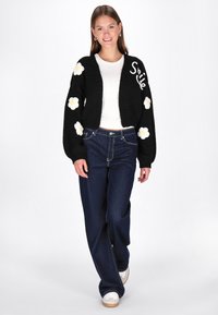 Juodas megztinis su baltais siuviniais gėlėmis, tekstu "Šypsena" ir grublėtomis rankovėmis; dėvimas ant balto marškinėlio ir priderintas prie tamsių džinsų.