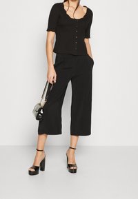 Haut noir côtelé à manches courtes avec encolure festonnée, assorti à un pantalon noir à jambes larges. Sandales noires à talons épais et petit sac.