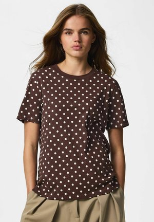 Jeune femme aux longs cheveux bruns portant un t-shirt marron à manches courtes avec des pois blancs et un pantalon beige sur un fond uni.