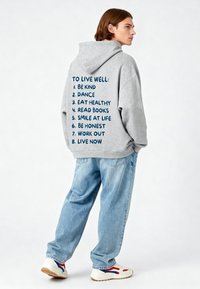 Felpa grigia dal taglio ampio, caratterizzata da un elenco inciso di suggerimenti per il benessere in blu navy sul retro. Abbinata a jeans azzurri.
