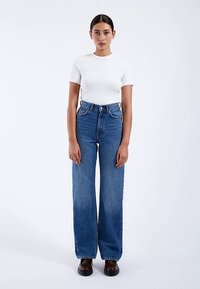 Jeans de mezclilla azul de talle alto con corte recto, que cuentan con bolsillos delanteros, trabillas para cinturón y un lavado ligeramente desteñido. Combinados con una camiseta blanca ajustada.