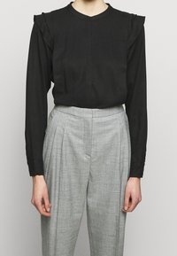 Blouse noire à manches longues avec épaules à volants, dotée d'un col rond et d'une fermeture à boutons sur le devant, associée à un pantalon gris plissé.