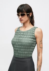 Gewatteerd groen mouwloos topje met een getextureerd, doorgestikt ontwerp. Gecombineerd met een donkergrijze rok; model draagt slanke, ovale zonnebrillen.