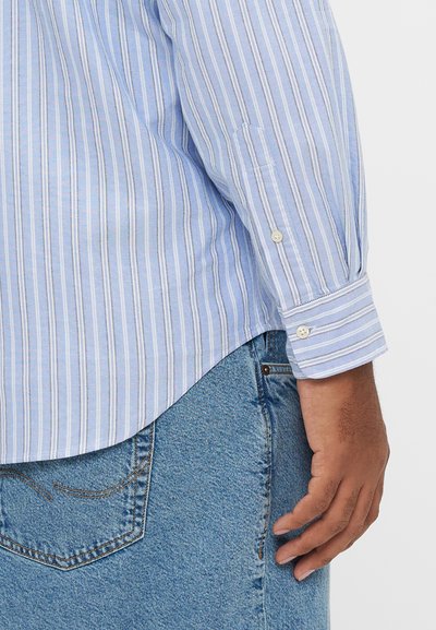 Camisa às riscas azul e branca de manga comprida com punhos abotoados, combinada com ganga azul clara, apresentando uma bainha curva e bolso lateral.