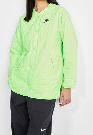Femme portant une veste matelassée Nike vert citron avec boutons-pression et un pantalon Nike noir avec logo blanc sur la jambe.