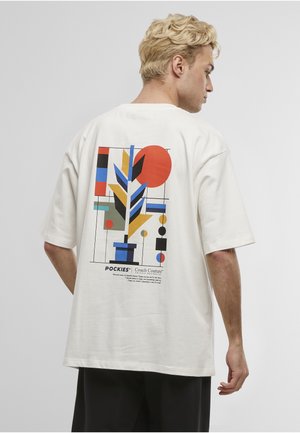 Blond mann med hvit t-skjorte med fargerikt abstrakt geometrisk plante-design og tekst på ryggen, sett fra siden.