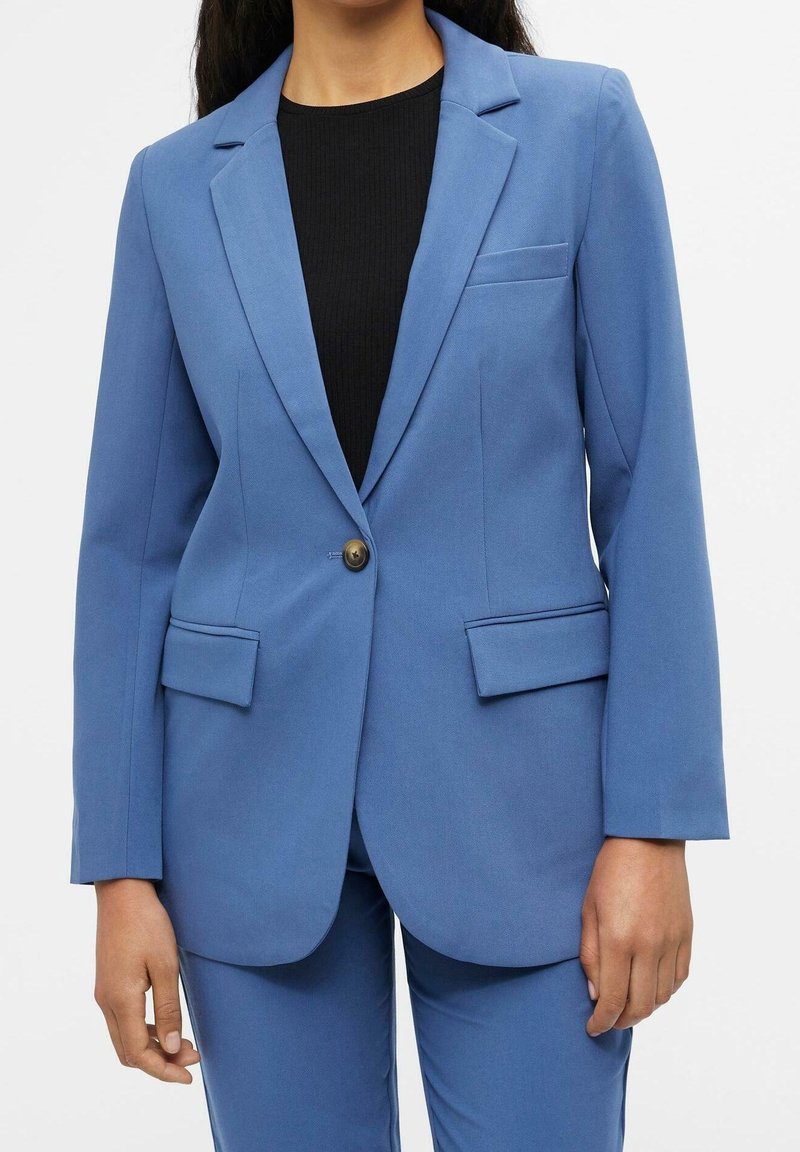 Femme portant un blazer bleu cintré avec un seul bouton, des poches à rabat, et un pantalon bleu assorti sur un top côtelé noir.