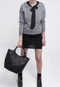 Sudadera gris con cuello y lazo negro, con ojales metálicos. Combinada con una falda negra, lleva un bolso tote negro texturizado. Botines negros.