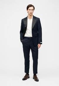 Bărbat purtând un costum bleumarin cu rever negru, cămașă albă și pantofi eleganți maro, stând cu o mână în buzunar pe un fundal alb.