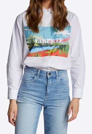 Person trägt ein weißes Hemd mit Knöpfen, das eine farbenfrohe Landschaft und den Schriftzug "LIBERTÉ" zeigt, kombiniert mit hellblauen, hoch taillierten Jeans.