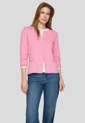 Frau trägt eine rosa Strickjacke über einem weißen Top und blauen Jeans und steht vor einem einfarbigen hellgrauen Hintergrund.