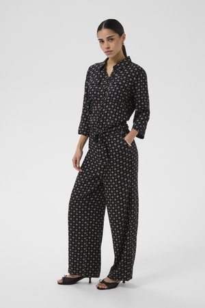 Žena s tmavými vlasmi v čiernom vzorovanom overal s gombíkmi a širokými nohavicami, jednou rukou v vrecku, nosiace čierne otvorené opätky.