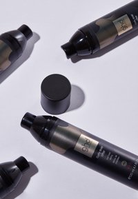 Trois flacons noires de spray de maintien de boucles ghd Curl Ever After, avec un bouchon retiré, disposés sur une surface claire.