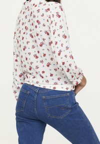 Blusa blanca ligera con estampado floral rojo y azul, combinada con jeans ajustados azul oscuro que tienen bolsillos traseros y detalles de costura en contraste.
