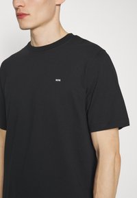 Černé bavlněné tričko s krátkým rukávem. Má malou bílou nášivku na hrudi. Jemná textura, klasický design s kulatým výstřihem.