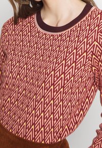 Strickpullover mit einem zigzagförmigen Muster in burgunderrot, pink und gelb. Verfügt über einen gerippten Kragen und lange Ärmel mit einer glatten Textur.