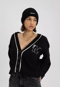 Czarny dziany kardigan z dekoltem w serek, białym wykończeniem i zapięciem na guziki, z wyszywanym literą "M". Połączony z czarną prążkowaną czapką.