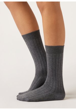 Beine mit dunkelgrauen, gerippten Socken, die bis zur Mitte der Wade reichen, stehen auf einer hellen Oberfläche vor einem schlichten Hintergrund.